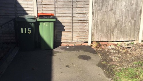 An outdoor scene showing two wheeled rubbish bins positioned against a wooden garden fence. The left bin is green and has the number '115' painted in white on its front, with an orange lid placed on top. The right bin is a darker shade of green, with no visible markings, and it leans slightly to the right. The ground beneath and around the bins is paved with a dark asphalt surface, with some patchy dark stains nearby. To the right of the bins, the fence extends upward and is constructed from vertical wooden planks, with a weathered appearance. The fence's lower edges are set on a narrow strip of soil and small plants that grow at the base. Bright daylight casts distinctive shadows on the pavement, creating areas of light and shadow on the ground. The scene reflects a typical residential or commercial waste disposal area, possibly in the Queens Park region, managed by Commercial Waste Queens Park, with the environment designed for rubbish collection and storage near a private or communal outdoor space.