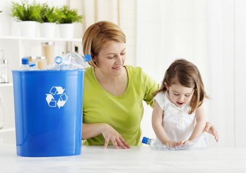 A woman with short, light brown hair wearing a bright yellow-green top is seated at a light-colored table, assisting a young girl with long brown hair dressed in a white top. The woman is smiling gently while guiding the girl as they handle a clear plastic bottle. To the left of them, a large blue recycling bin with a white recycling symbol on the front is positioned on the table, partially filled with crumpled and partly crushed plastic bottles. The background features a clean, bright room with white shelving units holding potted green plants and white containers, suggesting an organized indoor environment. The scene emphasizes responsible waste disposal and recycling practices, relevant to rubbish removal services in the local area such as Queens Park, highlighting an environmentally conscious approach to waste management.