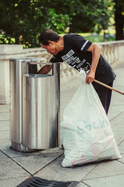 A person is seen disposing of paper waste into a small, light grey plastic bin with a black interior, situated on a wooden surface. The individual’s hand is placing a brown paper bag into the bin, w